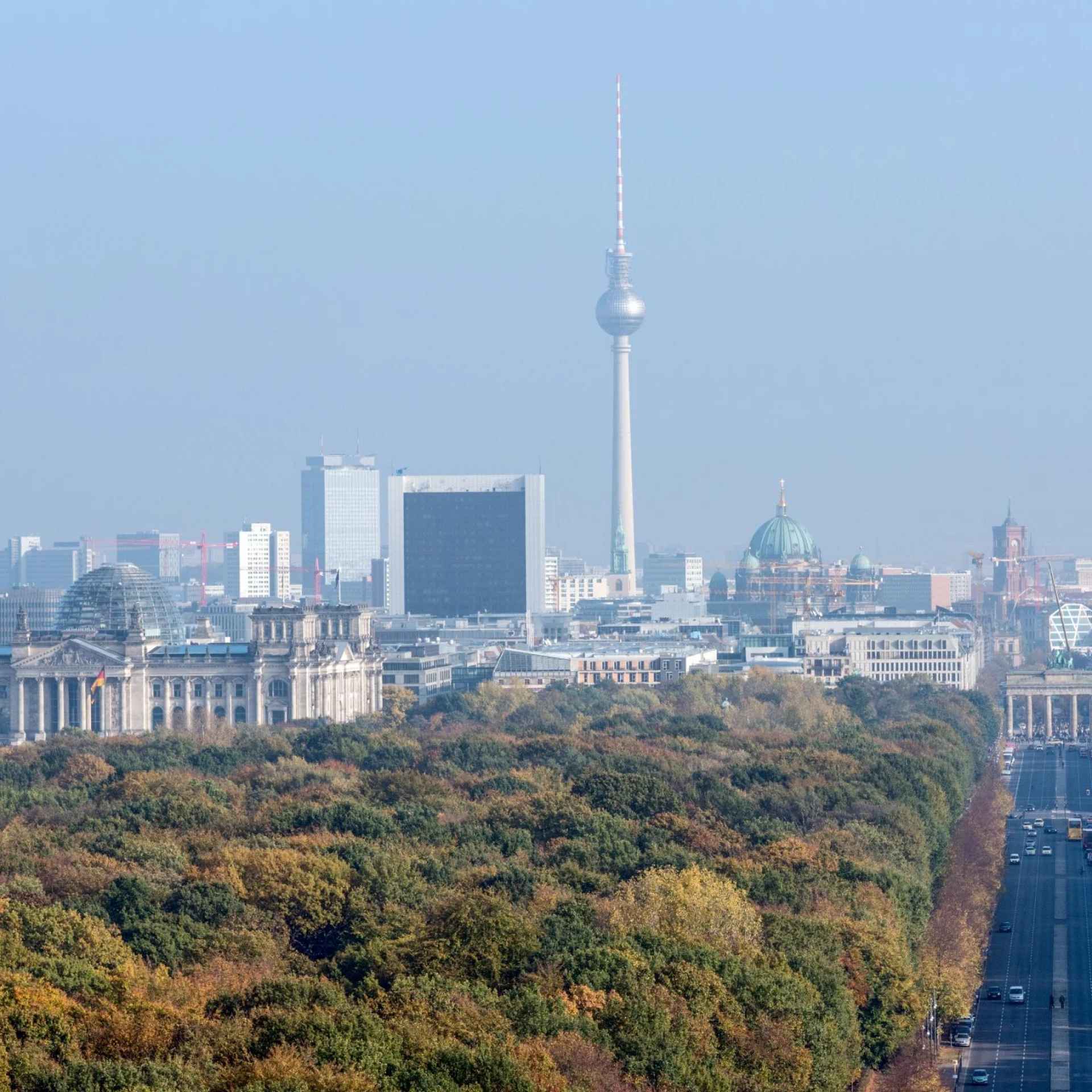 An aerial view of Berlin, Germany, showcasing the iconic landmarks such as the Reichstag building, Berlin TV Tower, and the Brandenburg Gate, surrounded by lush greenery and a clear sky.