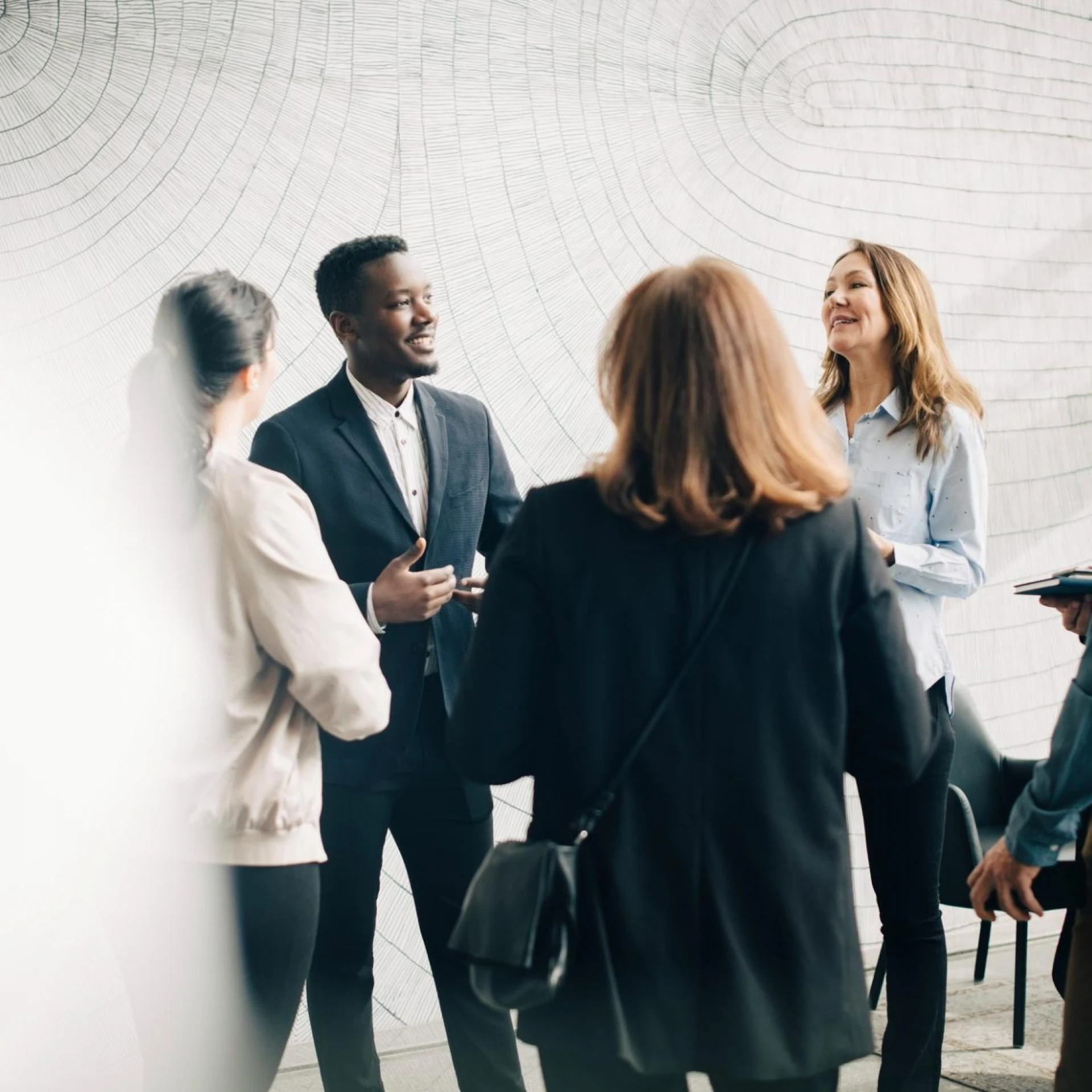 A group of professionals engaged in a lively discussion, standing together in a modern space with a textured wall in the background. Smiles and gestures suggest a positive and collaborative atmosphere.
