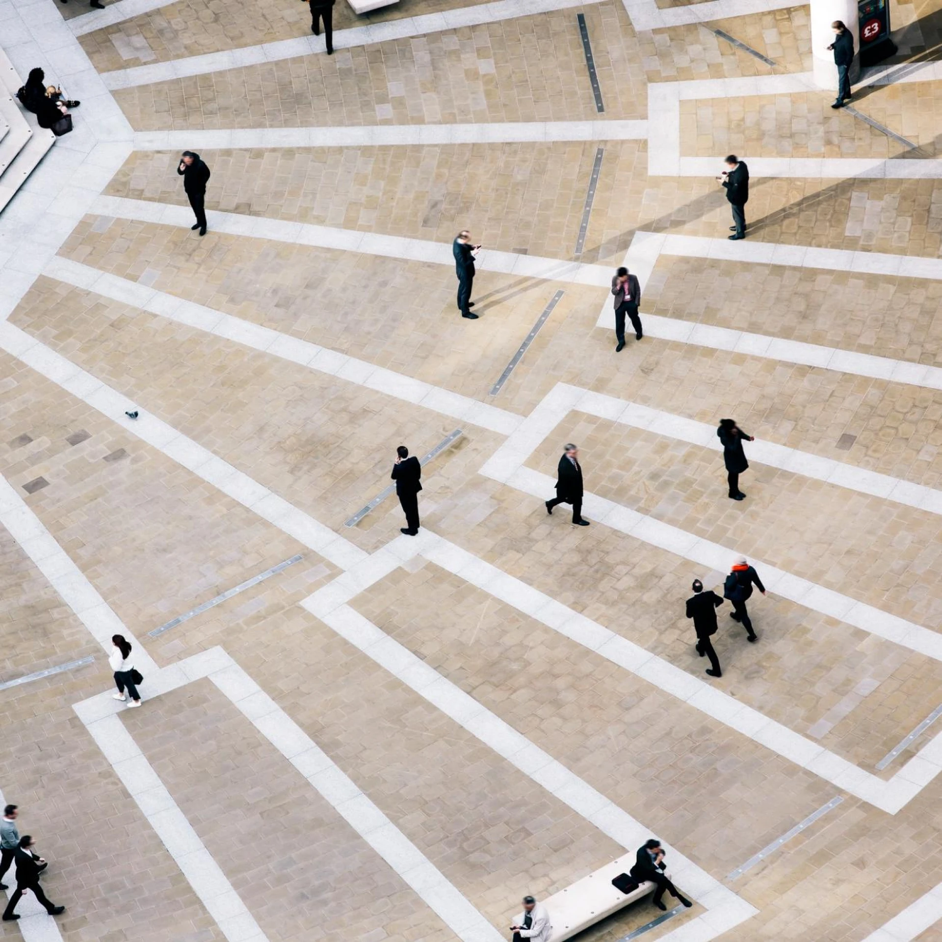 An aerial view of a public square with people walking and interacting. The geometric paving and strategic placement of individuals create a sense of movement and urban life.