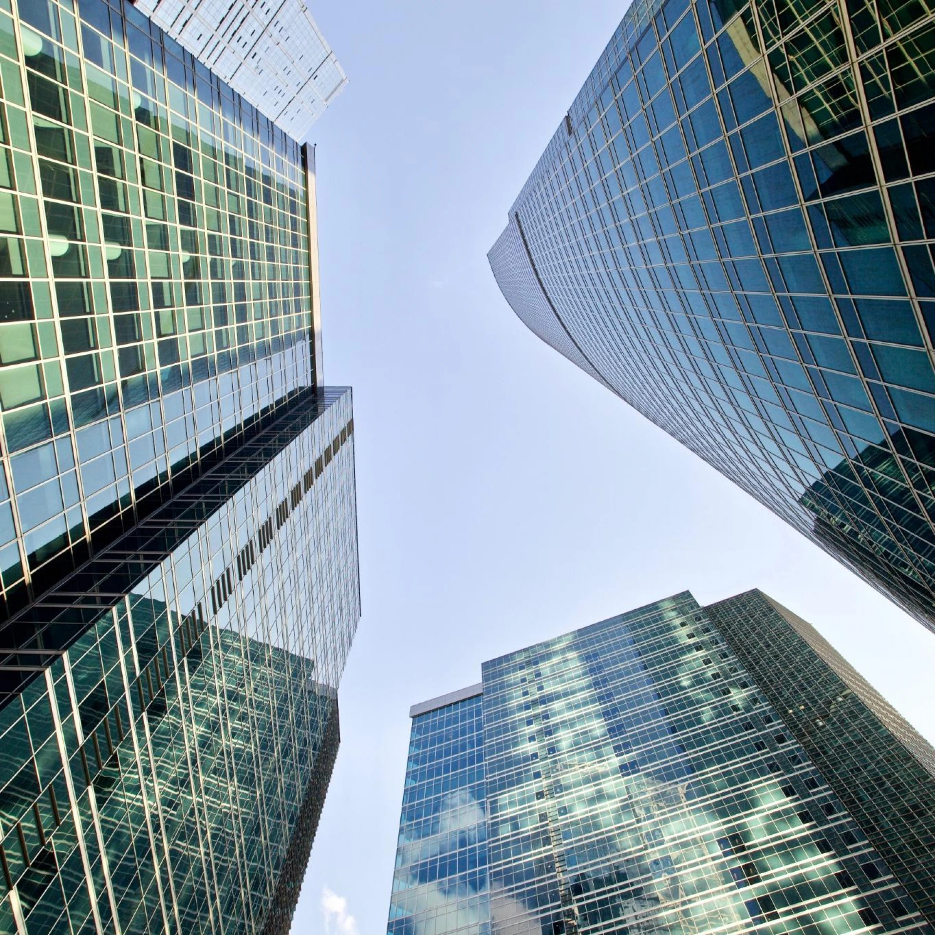 A low-angle view of modern skyscrapers with reflective glass facades against a clear blue sky. The architecture emphasizes urban sophistication and corporate ambition.