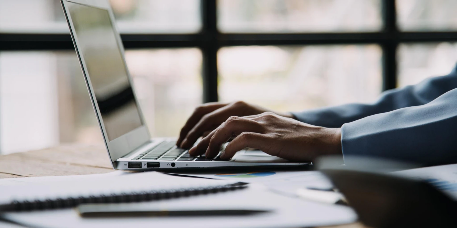 A person typing on a laptop placed on a wooden table, surrounded by documents, a pen, and a notebook. The background features a softly lit window, adding a calm and focused work setting.