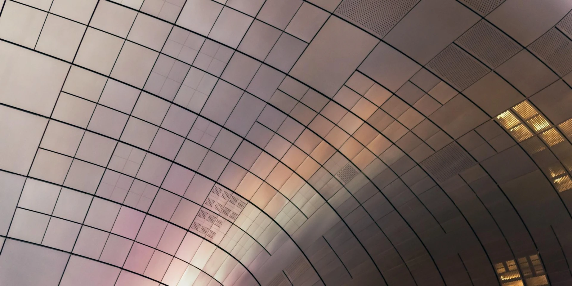 An abstract view of a curved ceiling with grid-like panels reflecting soft, gradient lighting. The design highlights modern architecture and innovative aesthetics.