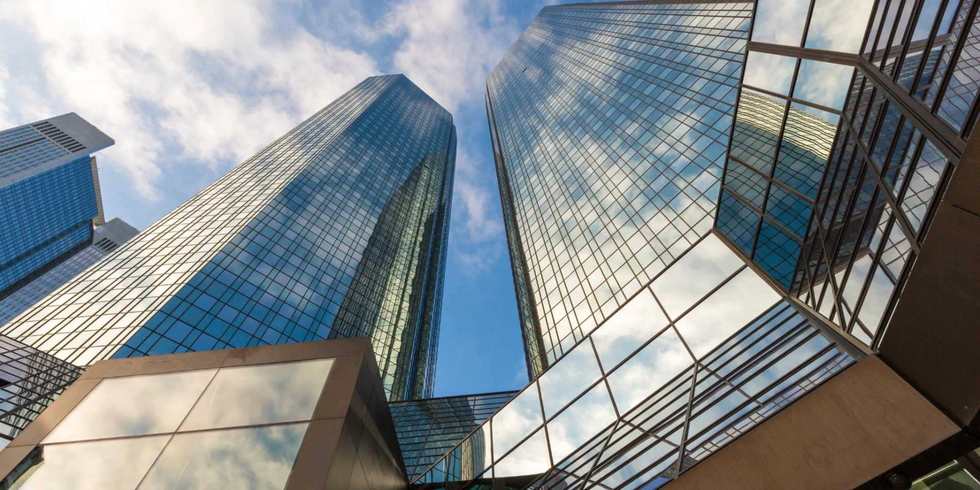 A low-angle view of tall, modern glass skyscrapers reflecting the blue sky and clouds. The geometric architecture emphasizes the sleek, urban design and professional corporate environment.