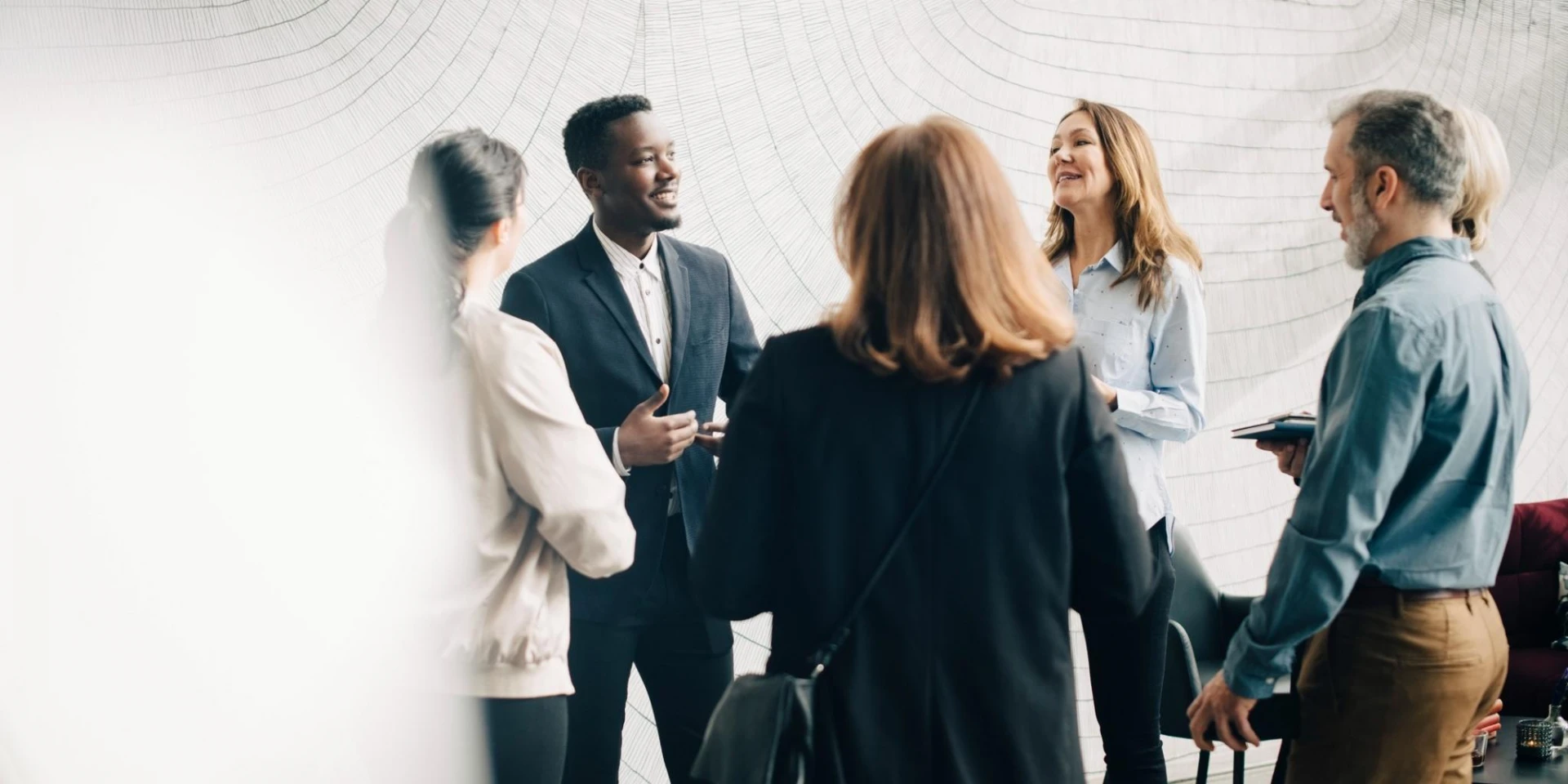 A group of professionals engaged in a lively discussion, standing together in a modern space with a textured wall in the background. Smiles and gestures suggest a positive and collaborative atmosphere.