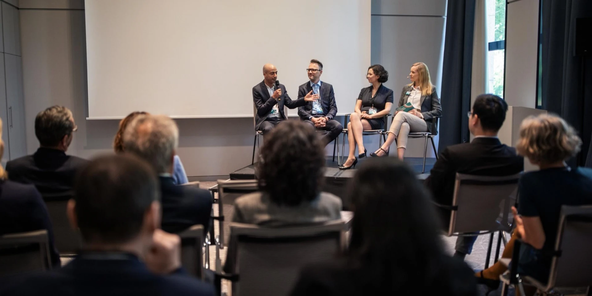 A panel of four speakers seated on stage, engaging with an audience in a conference room. One speaker is holding a microphone while others listen attentively.