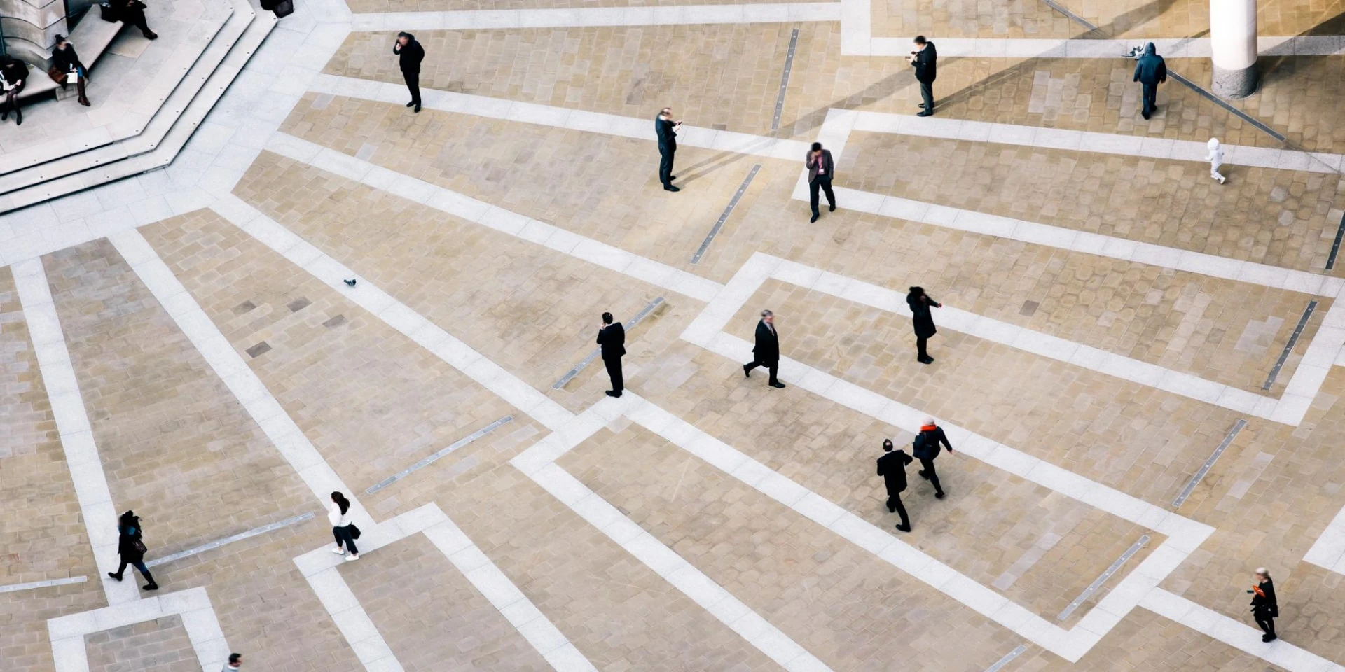 An aerial view of a public square with people walking and interacting. The geometric paving and strategic placement of individuals create a sense of movement and urban life.