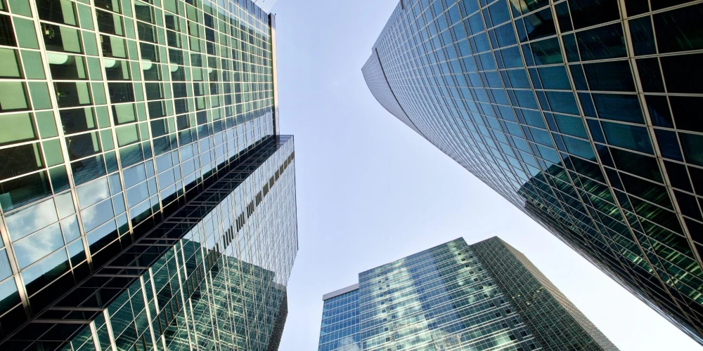 A low-angle view of modern skyscrapers with reflective glass facades against a clear blue sky. The architecture emphasizes urban sophistication and corporate ambition.