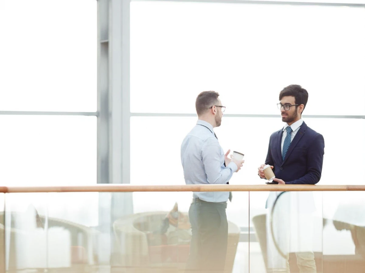 Two men in professional attire having a discussion in a bright, modern office space with large windows. Both are holding coffee cups, reflecting a casual yet professional environment.
