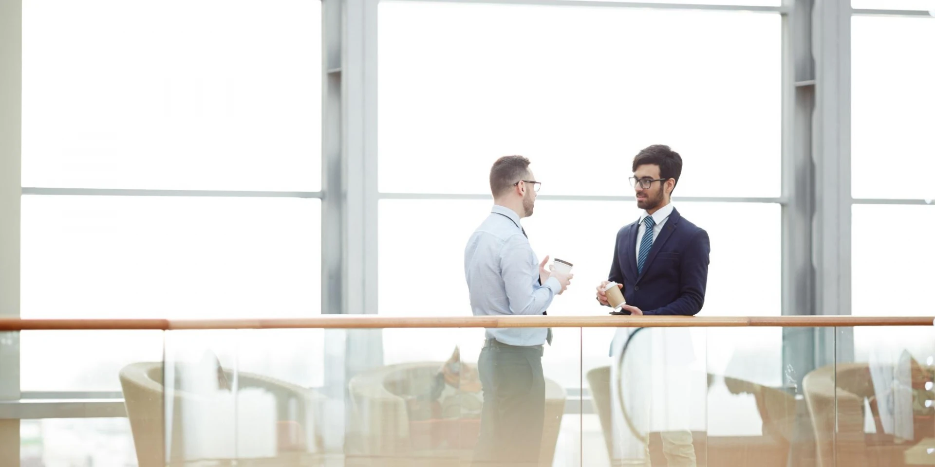 Two men in professional attire having a discussion in a bright, modern office space with large windows. Both are holding coffee cups, reflecting a casual yet professional environment.